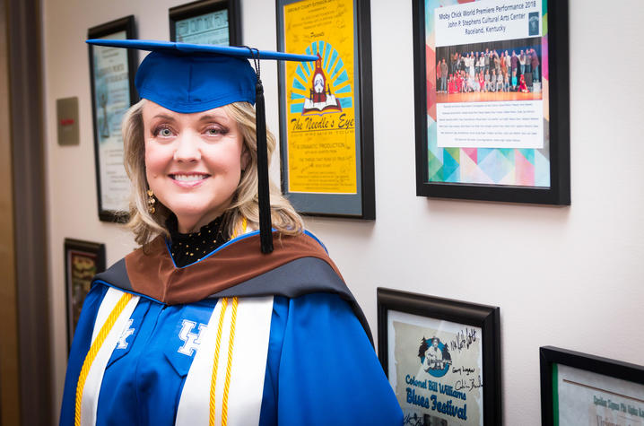 photo of Anne Stephens in her cap and gown with performance posters on wall behind her