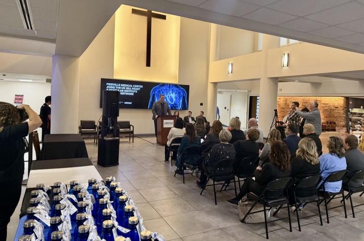 A speaker stands at a podium addressing an audience seated in an atrium during a formal event, with a presentation screen behind him and attendees listening and taking photos.