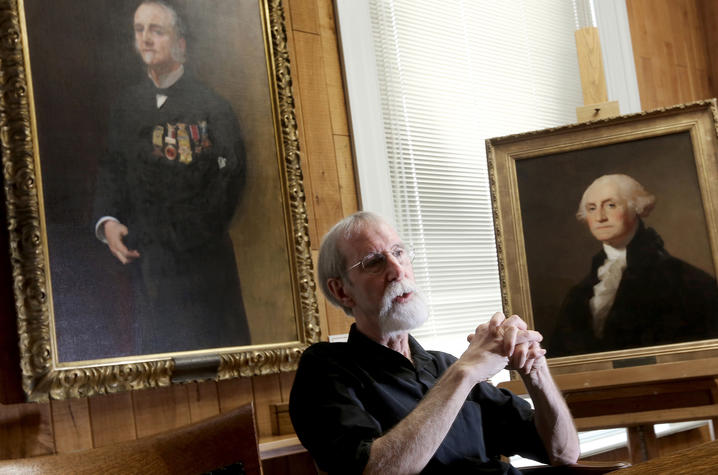 photo of Barry Bauman seated in front of portraits of Lucius Fairchild and George Washington by artists by John Singer Sargent and Thomas Sully