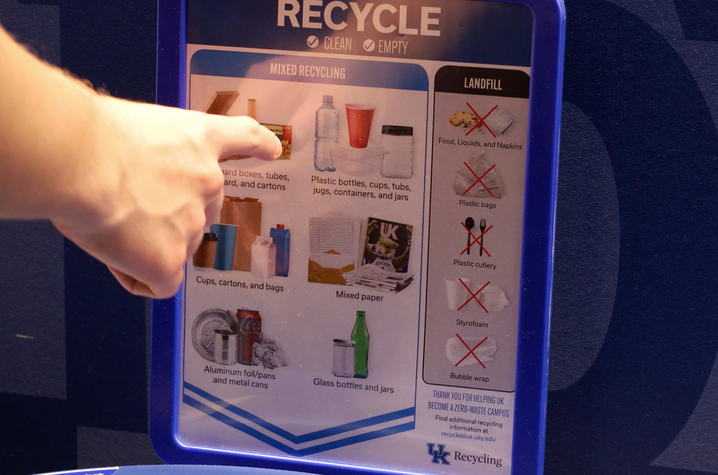 A person points to a recycling guide on a blue bin showing what items can and cannot be recycled at the University of Kentucky.