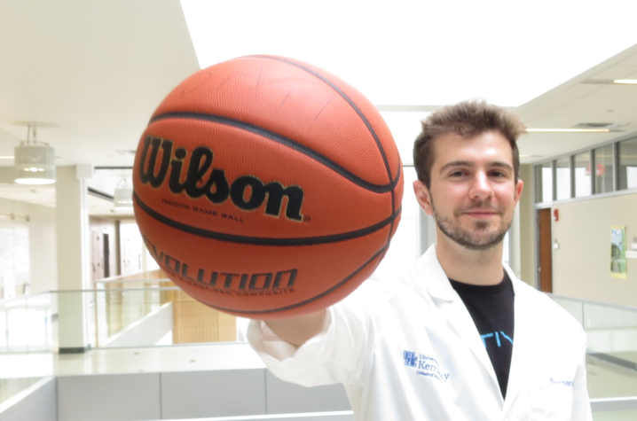 Man wearing a white lab coat holds a Wilson basketball prominently in front of them inside a bright, modern building.