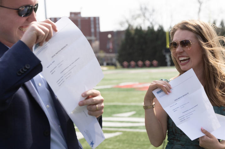 UK College of Medicine-Bowling Green Campus students Two people stand on an outdoor athletic field holding up printed documents, partially covering their upper bodies. A football field with yard lines, buildings and trees is visible in the background under bright daylight.