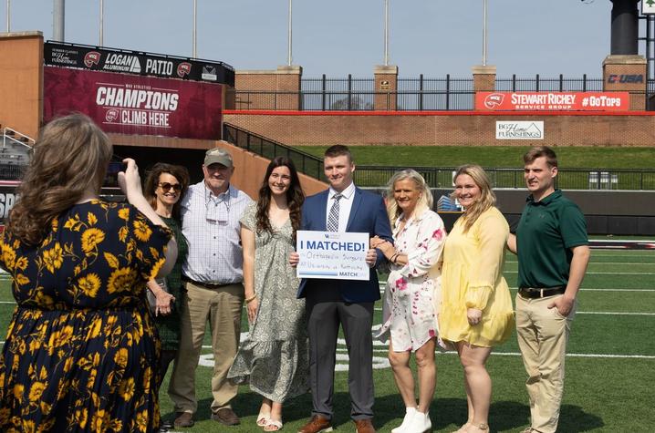 UK College of Medicine-Bowling Green Campus Match Day A group of people stands together on a football field, gathered around a person holding a sign that reads “MATCHED.” Stadium seating, a scoreboard with the word “Champions,” and campus buildings are visible in the background under bright daylight.