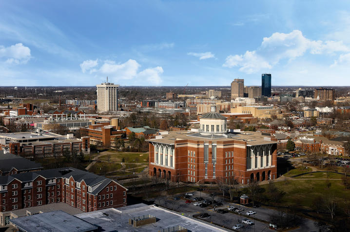 University of Kentucky campus Aerial view of the UK campus with a large red‑brick library building in the foreground, surrounding academic buildings, tree‑lined walkways, parking areas, and a distant city skyline under a blue sky with scattered clouds.