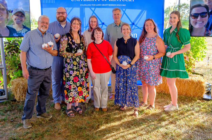Eight people standing in front of a blue “University of Kentucky” backdrop, holding small white objects.