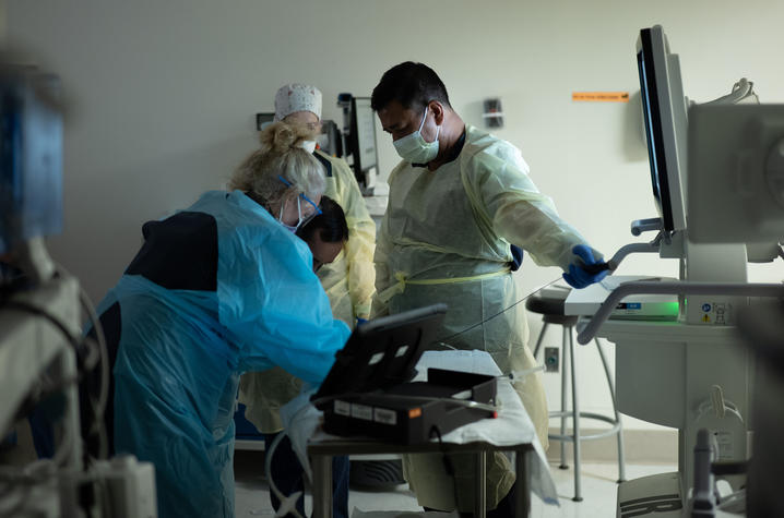 Clinical staff wearing protective gowns and masks work around a patient in a procedure room with medical equipment and monitors visible.
