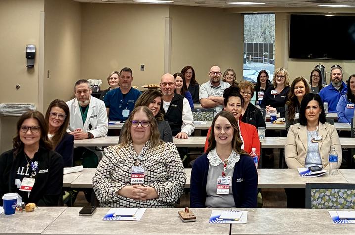 A large group of health care professionals sit at tables in a classroom-style setting, facing the camera and smiling during an orientation.