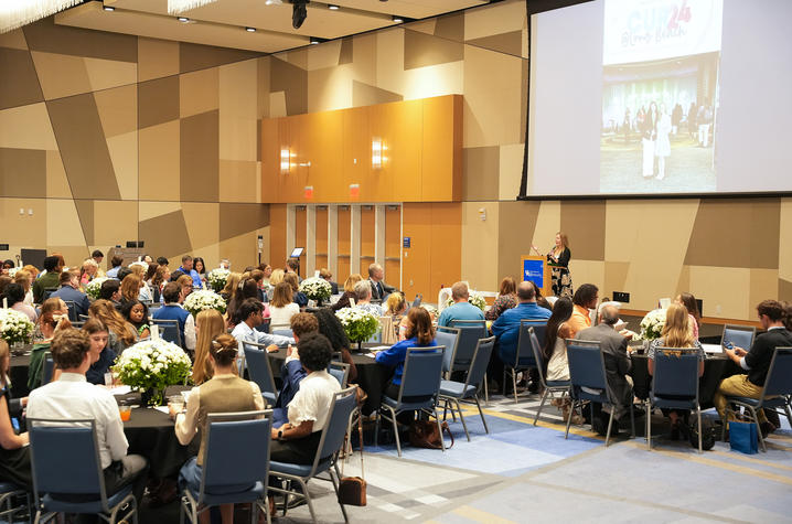 Attendees sit at tables in a large event space while a speaker presents from a podium at the front of the room.