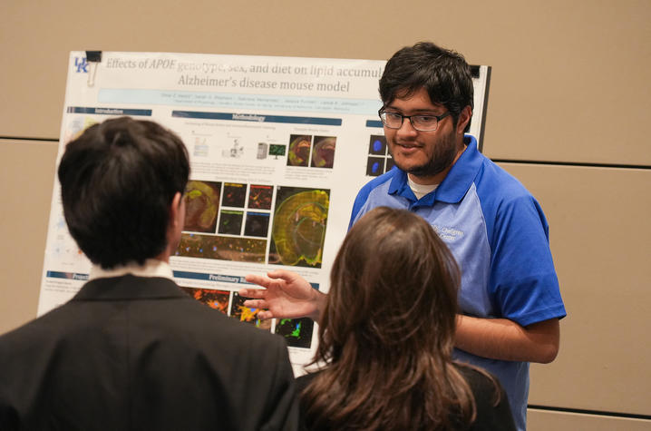 A person in a blue shirt presents a research poster to two attendees during an indoor event.