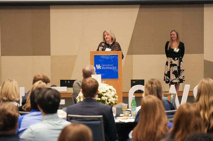 A speaker stands at a podium addressing an audience seated at tables, while another person stands nearby at the front of the room.