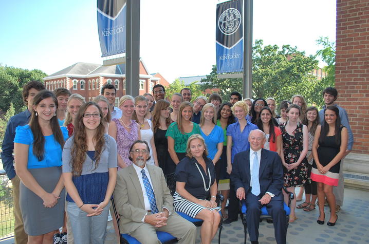A large group poses together outdoors in front of a brick building and campus banners.