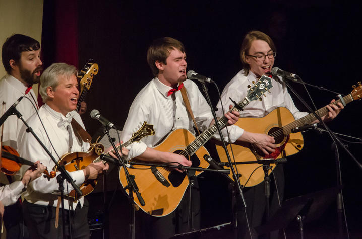 At left, Jefferson Johnson takes a break from directing to play his mandolin with UK’s bluegrass ensemble the Grass Cats, a popular ensemble at “Collage: A Holiday Spectacular.” Photo by Sally Horowitz Photography. A musical group of four men performing on stage, each wearing white shirts and bow ties; two are playing acoustic guitars, one is playing a violin, one is playing a mandolin, and all are singing into microphones under warm stage lighting.