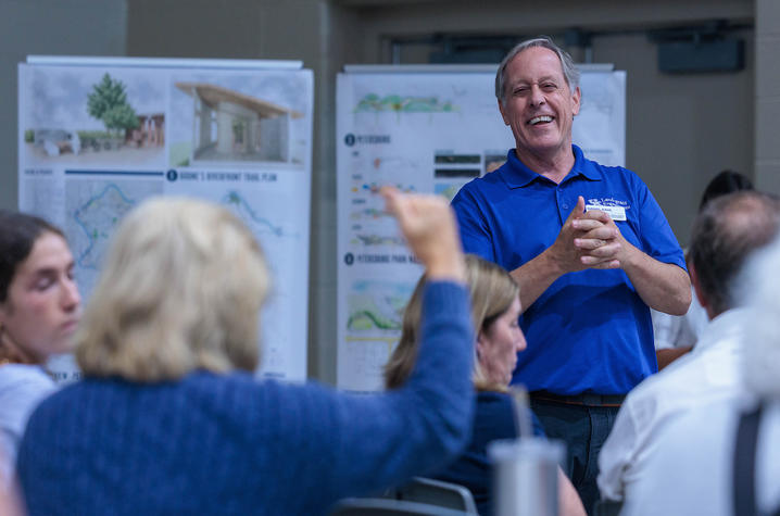A person in a blue polo shirt stands at the front of a community meeting or public workshop, speaking to attendees seated in the foreground. Large presentation boards displaying renderings, maps, and planning diagrams are positioned behind the speaker. 