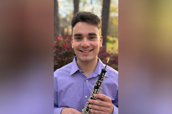 A person wearing a light purple button-down shirt holds an oboe upright while standing outdoors, with green foliage and pink flowers softly blurred in the background.