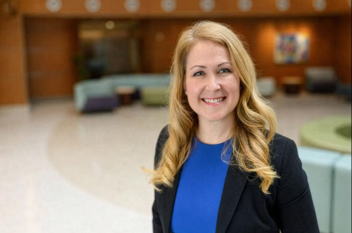 Photo of Lindsay Ragsdale A person with long, wavy blonde hair stands indoors in a spacious, modern lobby with soft seating, warm wood-paneled walls, and circular ceiling lights. The individual is wearing a blue top under a dark blazer.