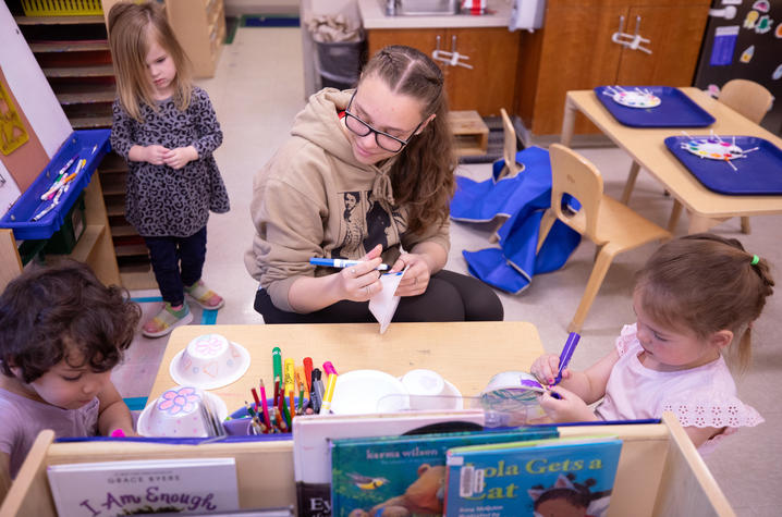 A teacher sits at a classroom table helping children decorate paper bowls with markers, while another child waits nearby in a colorful early learning classroom filled with art supplies and books.