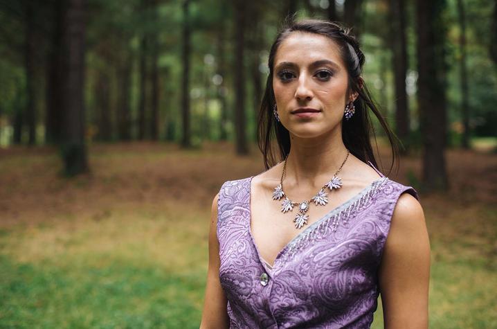 A woman with dark hair and a purple dress stands in the foreground with a forest behind her