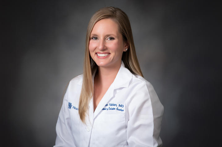 Photo of Denise Fabian Woman wearing a white medical lab coat with embroidered text on the chest, posed against a dark gray studio background.