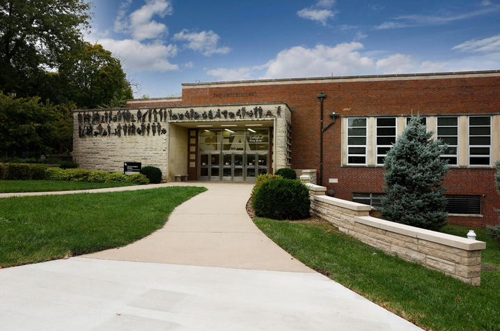 Brick academic building with a covered entrance, walkway and landscaping on a sunny day.