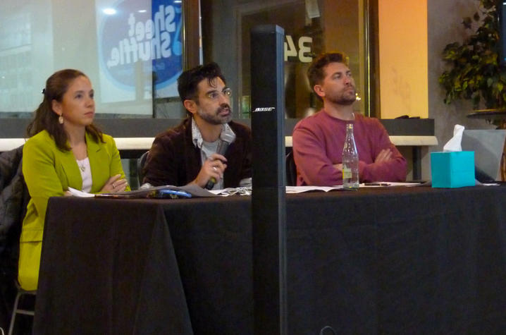 Three judges sit at a table watching a presentation, with papers, a microphone, and a tissue box in front of them. They appear attentive and focused, listening to a speaker off-camera.