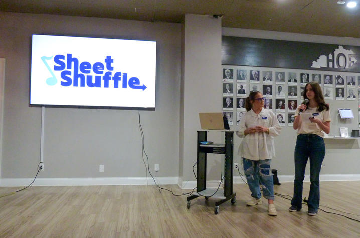 Two presenters stand at the front of a room giving a presentation beside a large screen displaying the logo “Sheet Shuffle.” One person speaks into a microphone while the other stands near a laptop on a cart. A photo wall is visible in the background.