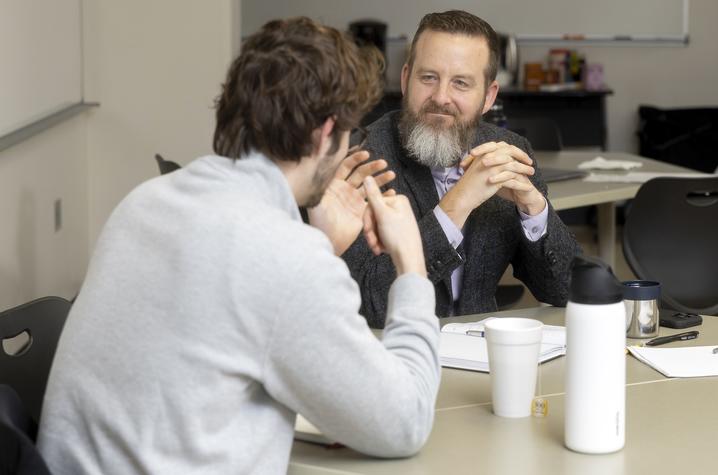 photo of Professor Lou Hirsch with student Two people sit across from each other at a table in a meeting room, engaged in conversation. Papers, notebooks and a coffee cup are on the table, and other tables and chairs are visible in the background.