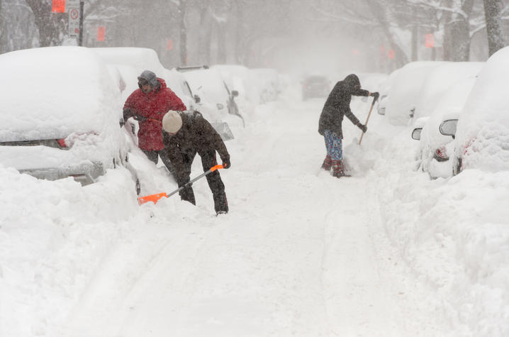 People shoveling snow on a street lined with cars during a heavy snowstorm.”