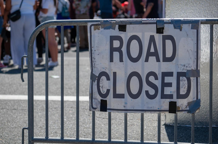 Metal barricades block a street with a white sign reading “ROAD CLOSED,” while pedestrians are visible in the background.