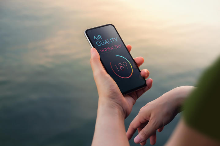 A person holds a smartphone displaying an air quality app with an “Unhealthy” alert and an air quality index reading of 189 against a blurred outdoor background.