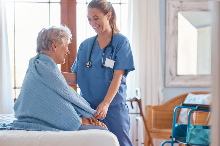 Health care professional in blue scrubs holding the hand of an older patient seated on a bed.