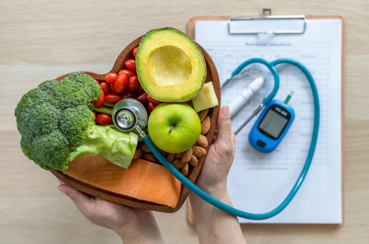 A pair of hands holds a heart-shaped bowl filled with healthy foods including broccoli, avocado, green apple, carrots, tomatoes, and nuts, next to a clipboard with a stethoscope and a blood glucose meter on a wooden surface.
