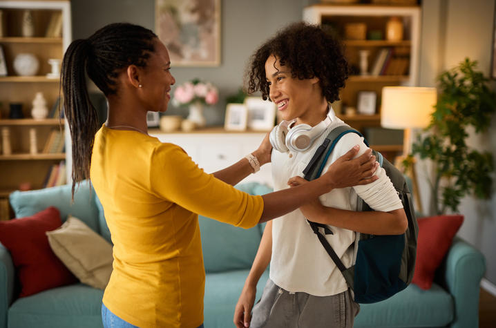 A young person wearing a backpack and headphones around their neck stands in a living room while another adult places their hands on the young person's shoulders. Bookshelves, a couch with pillows and a lamp are visible in the background.