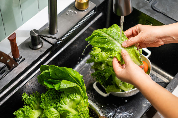 Hands rinse romaine lettuce under running water in a kitchen sink filled with fresh vegetables, including broccoli and leafy greens.