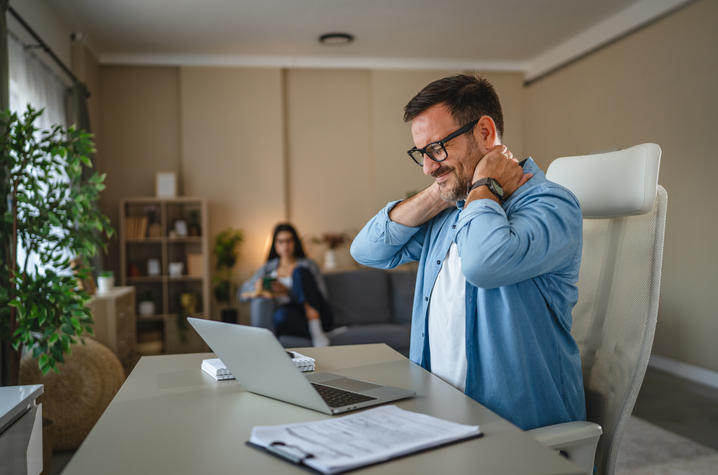 A man works on a laptop at a desk, rubbing his neck as if in discomfort, while another person sits blurred in the background of a home office.