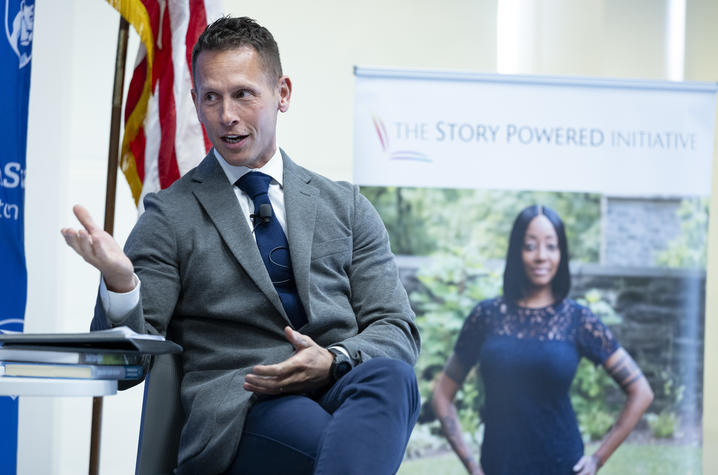 A panelist wearing a gray blazer and blue tie speaks while seated at an event, gesturing with one hand. 