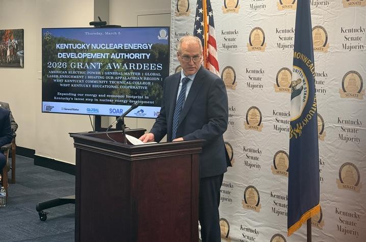 photo of KNEDA Grant Awardees Presentation A speaker stands at a wooden podium in a government meeting room, with U.S. and Kentucky flags behind them. A screen displays “Kentucky Nuclear Energy Development Authority 2026 Grant Awardees,” and a Kentucky Senate Majority backdrop fills the wall.