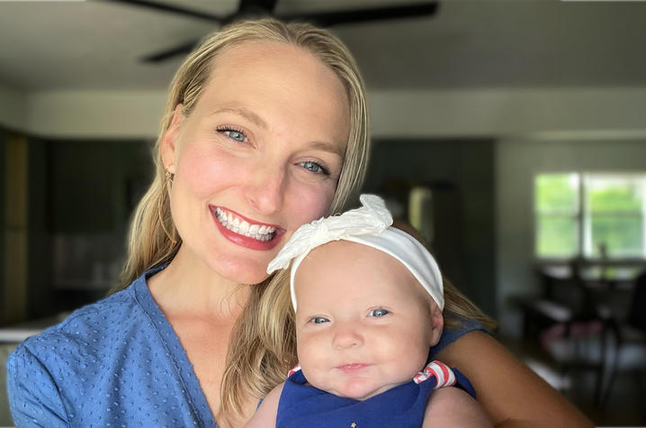 Close-up photo of an adult holding a baby wearing a white bow headband and blue outfit indoors, with a softly blurred home background.