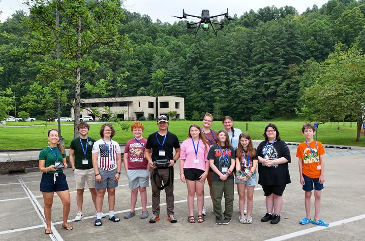 A group of students and instructors pose outdoors beneath a hovering drone. One instructor holds a drone controller while the participants stand together in a parking area surrounded by trees and a research facility.