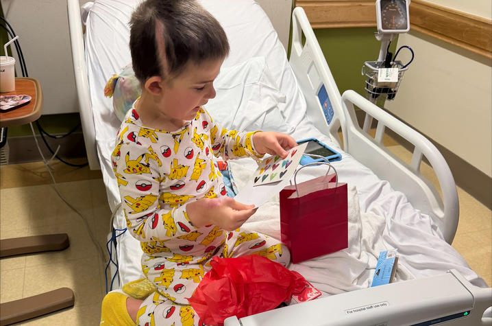 A young child wearing patterned pajamas sits upright in a hospital bed, opening a small red gift bag filled with Valentine-themed items. Medical equipment is visible around the bed, and the child appears focused on exploring the contents of the bag.