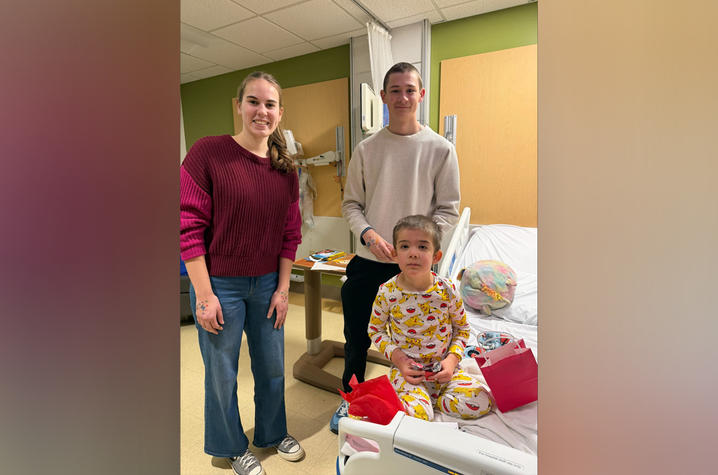 Two adults stand beside a child who is sitting on a hospital bed. The child is smiling and holding craft supplies, with a red gift bag in front of them. The room has medical equipment and a green wall in the background.