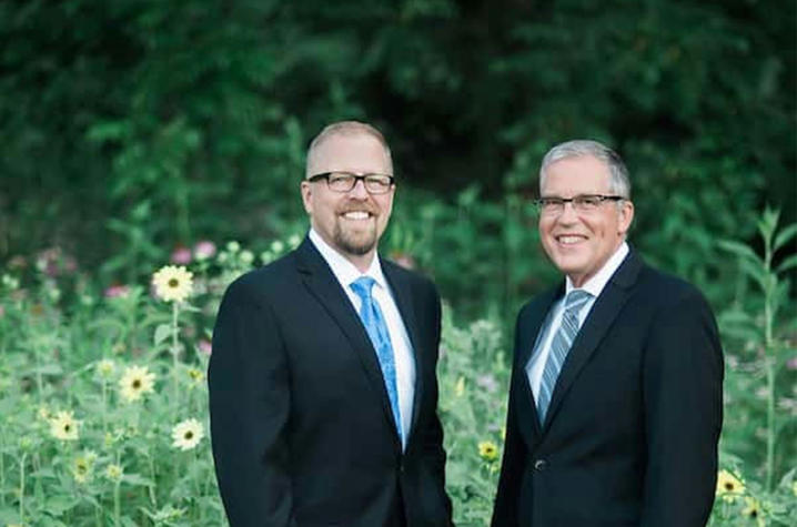 Photo of two white men wearing glasses and dark suits in front of a garden background