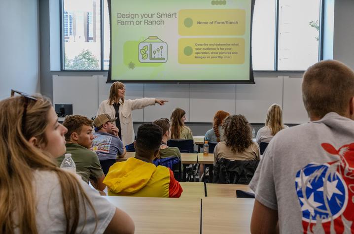 Participants sit at classroom-style tables during a workshop while a presenter stands at the front of the room pointing toward attendees. A large projection screen displays a slide titled “Design your Smart Farm or Ranch,” with prompts for naming a farm.