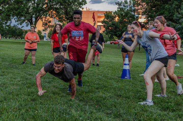 Participants take part in an outdoor team competition on a grassy field near campus buildings at dusk. One participant in a black shirt crawls toward a marker cone while another participant in a red shirt runs closely behind. 