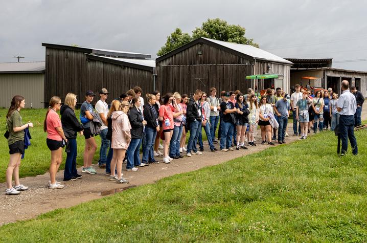 A large group of participants wearing name badges stands along a gravel path during a farm tour, listening to a speaker near several agricultural buildings. Green grass surrounds the path, and farm equipment is visible in the background.