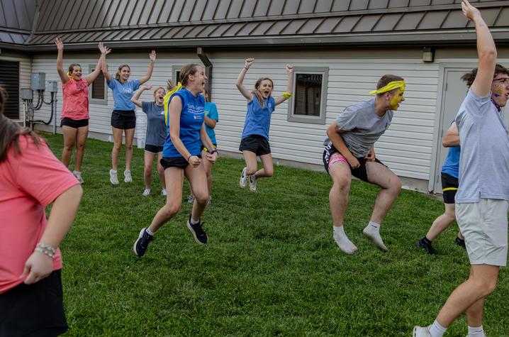 Participants in matching team colors take part in an outdoor group activity on a grassy lawn beside a building. Several participants wearing yellow bandanas and face paint jump into the air with raised arms while others run across the field. 