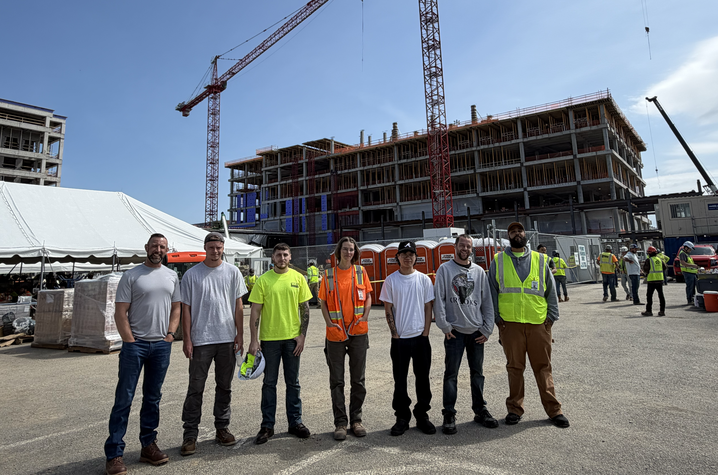 A group of construction workers stand together on a job site with a partially built structure and crane in the background.