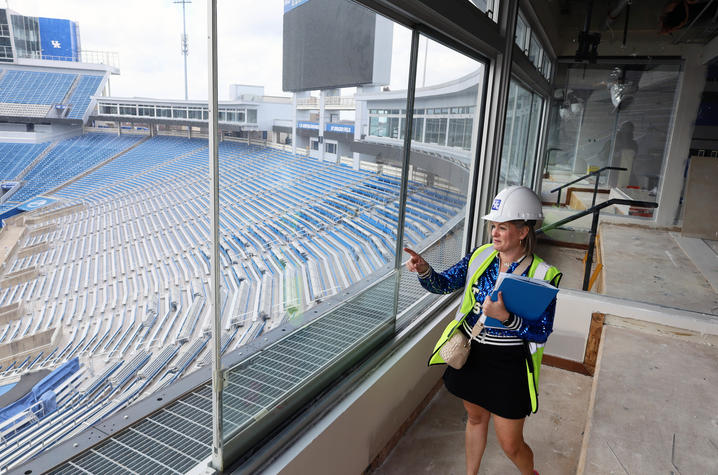 A woman wearing a hard hat and safety vest stands inside a stadium suite under construction, pointing out the window toward the seating bowl while holding a blue folder.