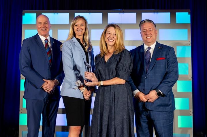 Photo of EmPATH leadership with award Four people stand on a stage in front of a blue-lit backdrop composed of rectangular light panels. Two individuals in the center hold a glass or crystal award together, positioned at chest height, while the other two stand on either side.