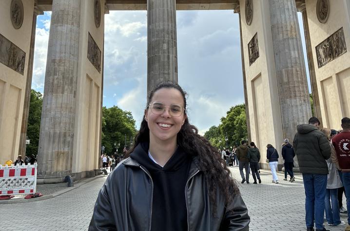Person standing in front of the Brandenburg Gate in Berlin, Germany, with the landmark’s columns and the Quadriga sculpture visible overhead. Pedestrians and an overcast sky appear in the background.