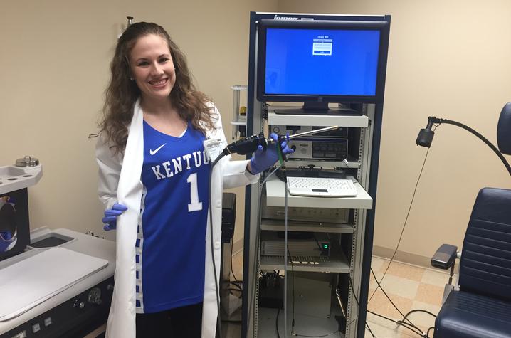 Rachel in a blue Kentucky basketball jersey with a white lab coat over it stands holding a rigid endoscope in her left hand